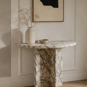 May include: A semi-circular console table crafted from white marble with grey veining, featuring a cross-shaped pedestal base. On the tabletop sits a matte beige ceramic vase containing dried baby's breath flowers and a small, shallow beige bowl.