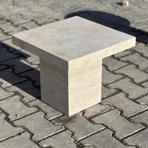 Può includere: Un tavolino da salotto quadrato in pietra beige chiaro con un piano spesso e testurizzato e una base quadrata. Il tavolino è posizionato su un patio in mattoni grigi.