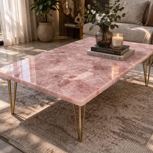 May include: A rectangular coffee table with a pink, patterned surface and gold-colored hairpin legs. The table is in a living room setting, with books, a candle, and a vase on top. The table's dimensions are approximately 48 inches long and 24 inches wide.