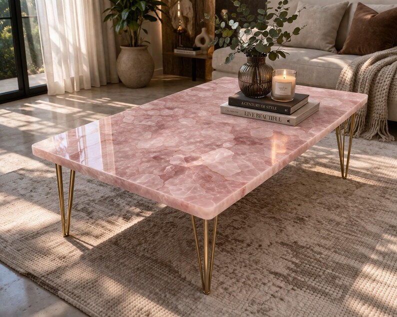 May include: A rectangular coffee table with a pink, patterned surface and gold-colored hairpin legs. The table is in a living room setting, with books, a candle, and a vase on top. The table's dimensions are approximately 48 inches long and 24 inches wide.