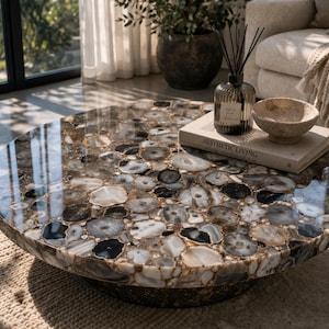 May include: A circular coffee table with a polished agate stone surface in grey, white, and brown tones. The table has a dark base and sits on a woven rug. A book, bowl, and diffuser are on the table.