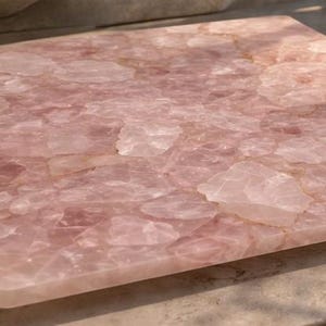 May include: A square, pink quartz serving board with a mosaic-like pattern. The board has a polished surface and is set on a light-colored countertop. The quartz has a translucent quality, with variations in color and texture.