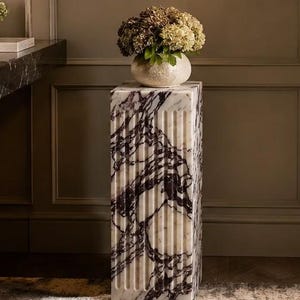May include: A rectangular marble pedestal featuring a fluted design with deep purple and black veining against a white stone base. The column has a smooth top surface supporting a round ceramic vase filled with a cluster of dried hydrangea flowers.