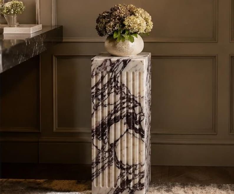 May include: A rectangular marble pedestal featuring a fluted design with deep purple and black veining against a white stone base. The column has a smooth top surface supporting a round ceramic vase filled with a cluster of dried hydrangea flowers.