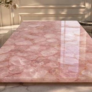 May include: A rectangular, polished rose quartz slab. The pink stone has a glossy finish and a pattern of irregular shapes. The slab is likely used as a decorative surface or serving board. A candle and books are in the background.