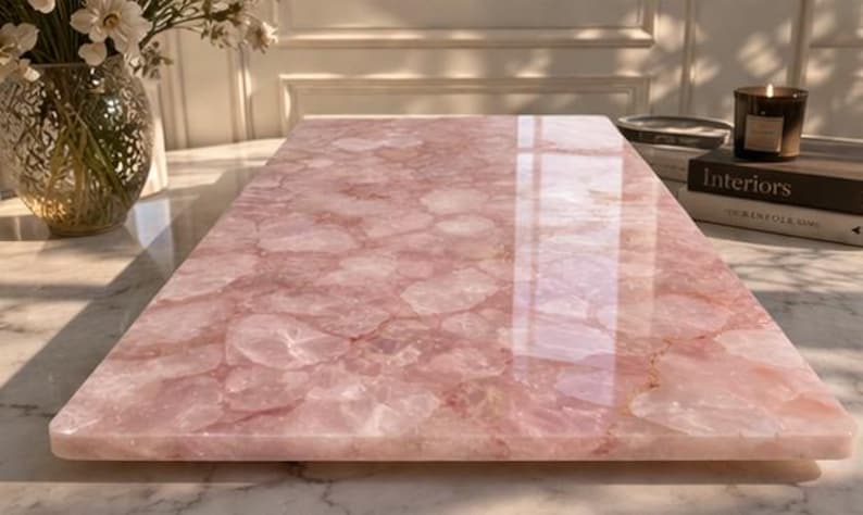 May include: A rectangular, polished rose quartz slab. The pink stone has a glossy finish and a pattern of irregular shapes. The slab is likely used as a decorative surface or serving board. A candle and books are in the background.