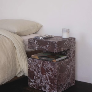 Può includere: Un comodino moderno effetto marmo, con un motivo rosso scuro e bianco. Il comodino ha due livelli, con un libro e un vassoio decorativo sopra. Una candela è sul livello superiore. Il comodino è accanto a un letto con biancheria da letto beige.