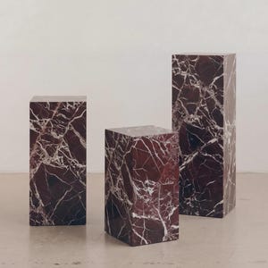 May include: Three rectangular pedestals with a rich burgundy marble appearance and white veining. The pedestals vary in height, creating a visually interesting display. The marble has a polished finish, reflecting light and adding a touch of luxury to the setting.