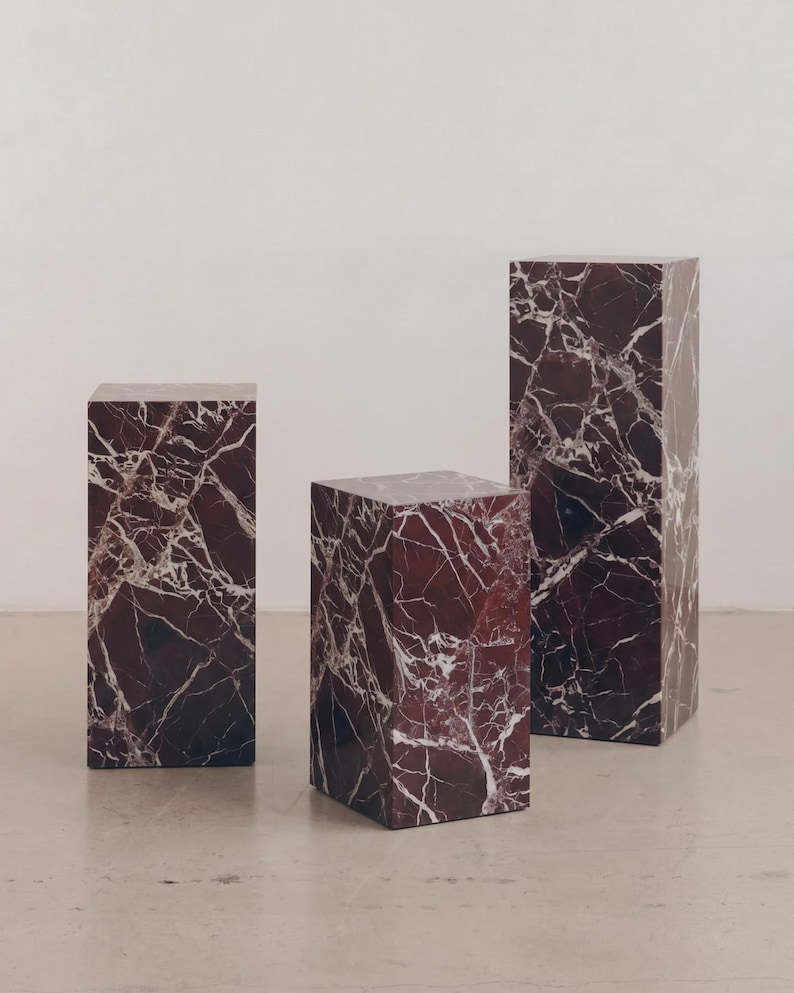 May include: Three rectangular pedestals with a rich burgundy marble appearance and white veining. The pedestals vary in height, creating a visually interesting display. The marble has a polished finish, reflecting light and adding a touch of luxury to the setting.