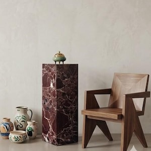 May include: A wooden chair with geometric design and a tall, rectangular marble pedestal with a small green and gold decorative object on top. Several ceramic vases with painted designs are on the left.