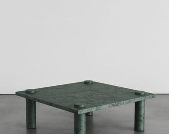 Puede incluir: Una mesa de centro cuadrada fabricada en mármol verde oscuro con un patrón de vetas naturales visible. La mesa presenta una superficie superior plana sostenida por cuatro patas cilíndricas gruesas situadas en cada esquina, con detalles circulares.