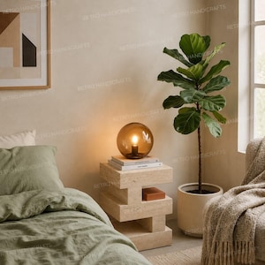 May include: A spherical amber glass table lamp sits atop a stacked beige travertine stone side table. The lamp emits a warm glow. Beside the table is a tall fiddle leaf fig plant in a white pot, and a sage green linen bed set is visible.