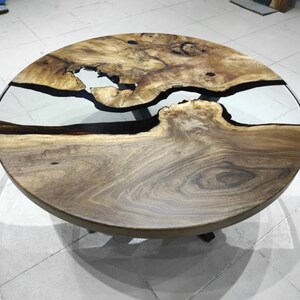May include: Circular wooden coffee table with a natural wood grain. It features a clear epoxy resin river design, creating a unique visual effect. The table is supported by black metal legs. The table's diameter is approximately 100 cm.