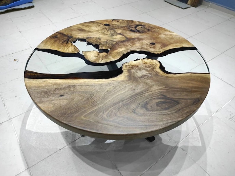 May include: Circular wooden coffee table with a natural wood grain. It features a clear epoxy resin river design, creating a unique visual effect. The table is supported by black metal legs. The table's diameter is approximately 100 cm.