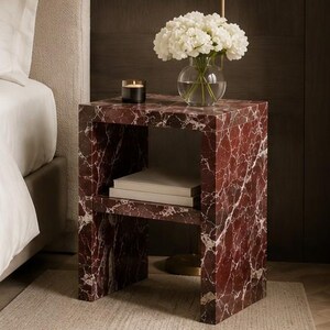 May include: A marble-effect bedside table in burgundy and white. The open shelf holds books, and the top displays a vase of white flowers and a candle. The table is next to a bed. The table is made of stone.
