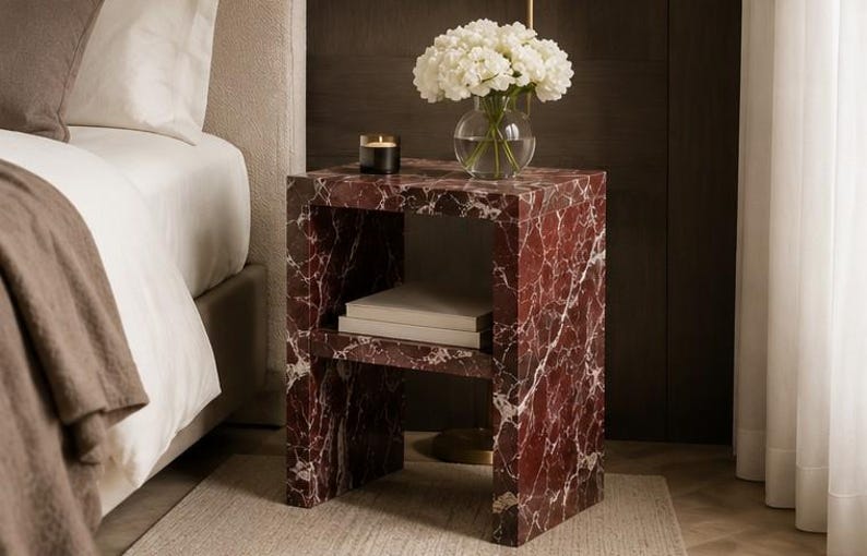 May include: A marble-effect bedside table in burgundy and white. The open shelf holds books, and the top displays a vase of white flowers and a candle. The table is next to a bed. The table is made of stone.