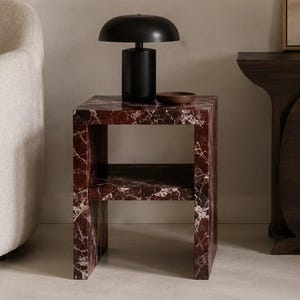 May include: A contemporary side table with a red marble effect and a black lamp. The table has a shelf and a small wooden bowl. The lamp has a rounded shade and a cylindrical base. The table is next to a cream armchair.