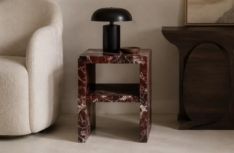 May include: A contemporary side table with a red marble effect and a black lamp. The table has a shelf and a small wooden bowl. The lamp has a rounded shade and a cylindrical base. The table is next to a cream armchair.