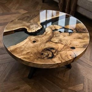 May include: A round coffee table with a unique wood grain design and a glossy, clear resin finish. The table has a dark, natural wood colour with black legs. The table's diameter is approximately 100 cm.