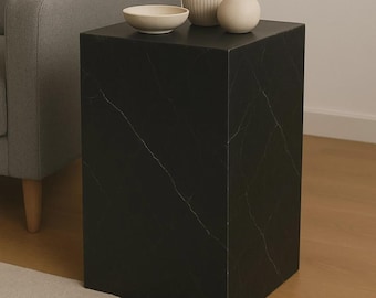 May include: A black marble-look side table with a square shape. The table has white veining and is topped with a small white bowl, a ribbed vase, and a round vase. The table is next to a gray armchair.