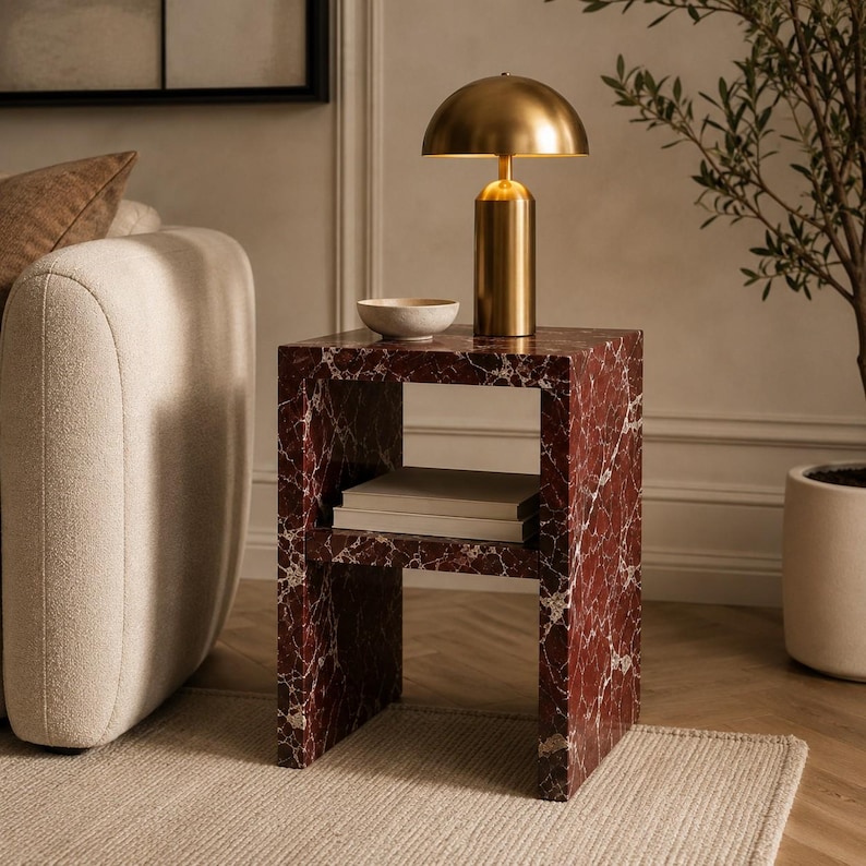 May include: A red marble side table with a gold lamp and a small bowl. The table has a shelf with books. The lamp has a rounded shade and a cylindrical base. The table is 51 cm tall.