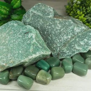 May include: A collection of green aventurine stones. The image features two large, rough-cut stones and several smaller, polished tumbled stones. The stones vary in shades of green, from light to dark, and are arranged on a light-colored surface.