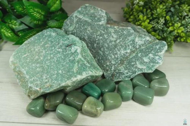 May include: A collection of green aventurine stones. The image features two large, rough-cut stones and several smaller, polished tumbled stones. The stones vary in shades of green, from light to dark, and are arranged on a light-colored surface.