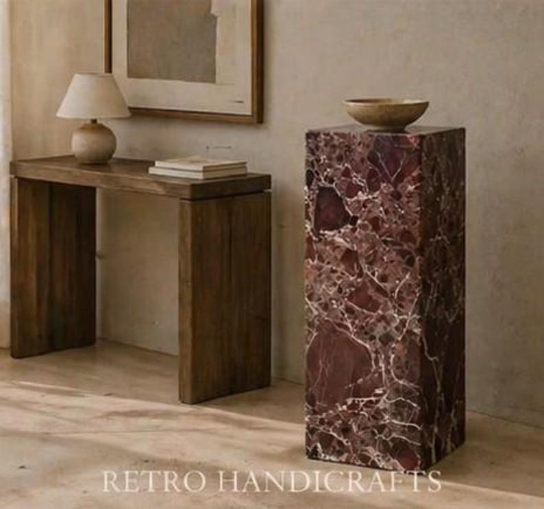 May include: A dark wooden console table with a small lamp and books, and a tall, rectangular marble pedestal with a bowl on top. The pedestal is a deep red color with white veining. The text "RETRO HANDICRAFTS" is at the bottom.