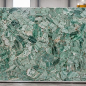 May include: Large rectangular slab of green stone with a mosaic-like pattern. The stone features varying shades of green and white, creating a textured appearance. The slab is displayed in a warehouse setting with other stone slabs in the background.