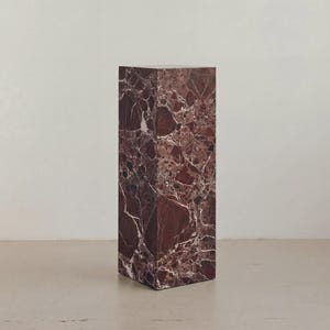 May include: A rectangular marble pedestal in shades of burgundy and white. The stone has a natural veining pattern. The pedestal is a solid block, suitable for displaying art or decor. The surface is smooth and polished.