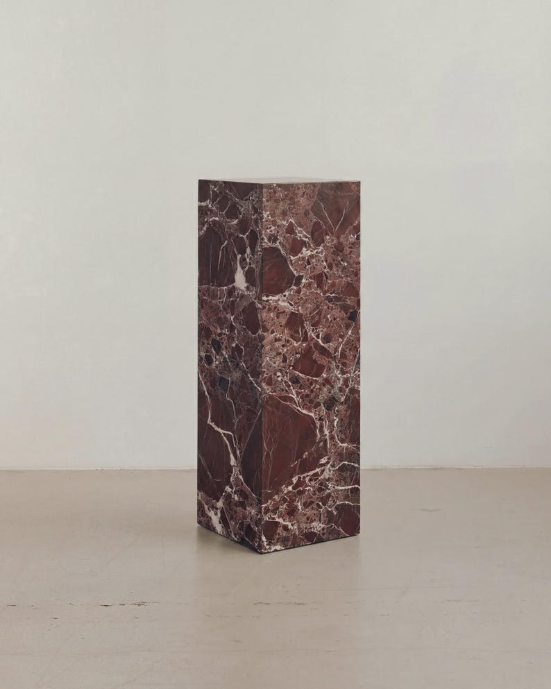 May include: A rectangular marble pedestal in shades of burgundy and white. The stone has a natural veining pattern. The pedestal is a solid block, suitable for displaying art or decor. The surface is smooth and polished.