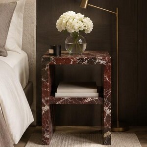 May include: A red and white marble-effect bedside table. A glass vase with white flowers and a black candle are on top. The table has a lower shelf with books. The table is approximately 50cm wide.