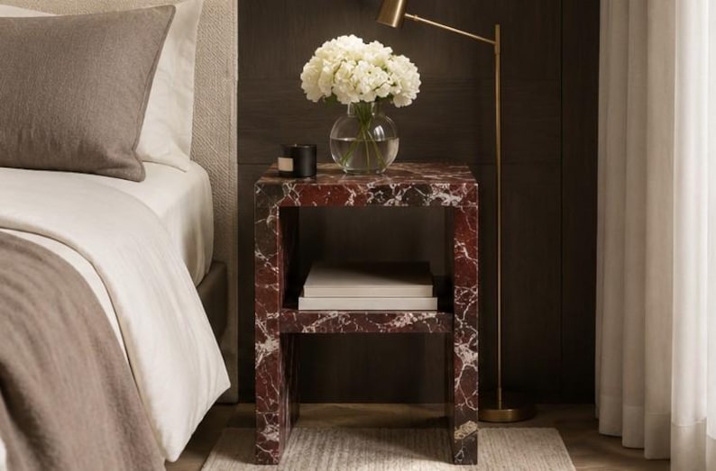 May include: A red and white marble-effect bedside table. A glass vase with white flowers and a black candle are on top. The table has a lower shelf with books. The table is approximately 50cm wide.