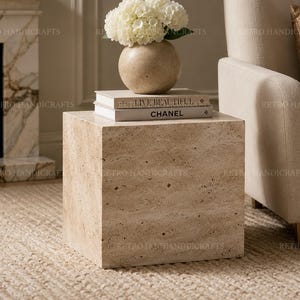 May include: A square, beige stone side table with a textured surface. A beige vase with white flowers sits on top, along with two books. A beige armchair is to the right, and a fireplace is in the background.