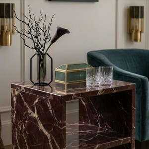 May include: A side table with a marble top featuring a dark red and white veined design. It displays a black metal and glass vase with branches and a dark purple calla lily, a green glass box, and two crystal glasses. A teal armchair is in the background.