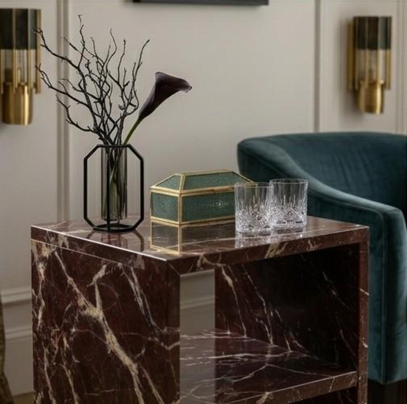 May include: A side table with a marble top featuring a dark red and white veined design. It displays a black metal and glass vase with branches and a dark purple calla lily, a green glass box, and two crystal glasses. A teal armchair is in the background.