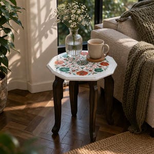 以下が含まれることがあります： 赤、緑、オレンジの花柄が施された白い天板の小さなサイドテーブル。テーブルは濃い茶色の曲線的な脚をしています。白い花が入ったガラスの花瓶とコーヒーカップが上に置かれています。テーブルの高さは約50cmです。