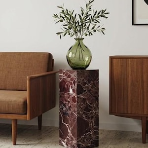 May include: A decorative display featuring a green glass vase with olive branches, placed on a rectangular, burgundy marble pedestal. A brown armchair and wooden cabinet are in the background. The pedestal has a unique marble pattern.