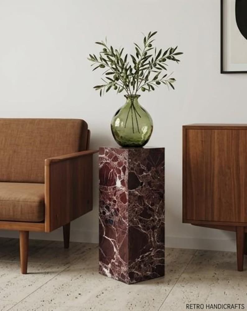 May include: A decorative display featuring a green glass vase with olive branches, placed on a rectangular, burgundy marble pedestal. A brown armchair and wooden cabinet are in the background. The pedestal has a unique marble pattern.