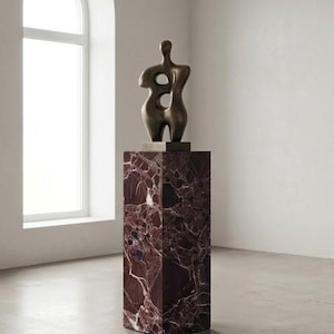 May include: A bronze sculpture of a human form with two large openings, mounted on a rectangular, dark red marble pedestal. The sculpture is in a bright, minimalist room with a large arched window.