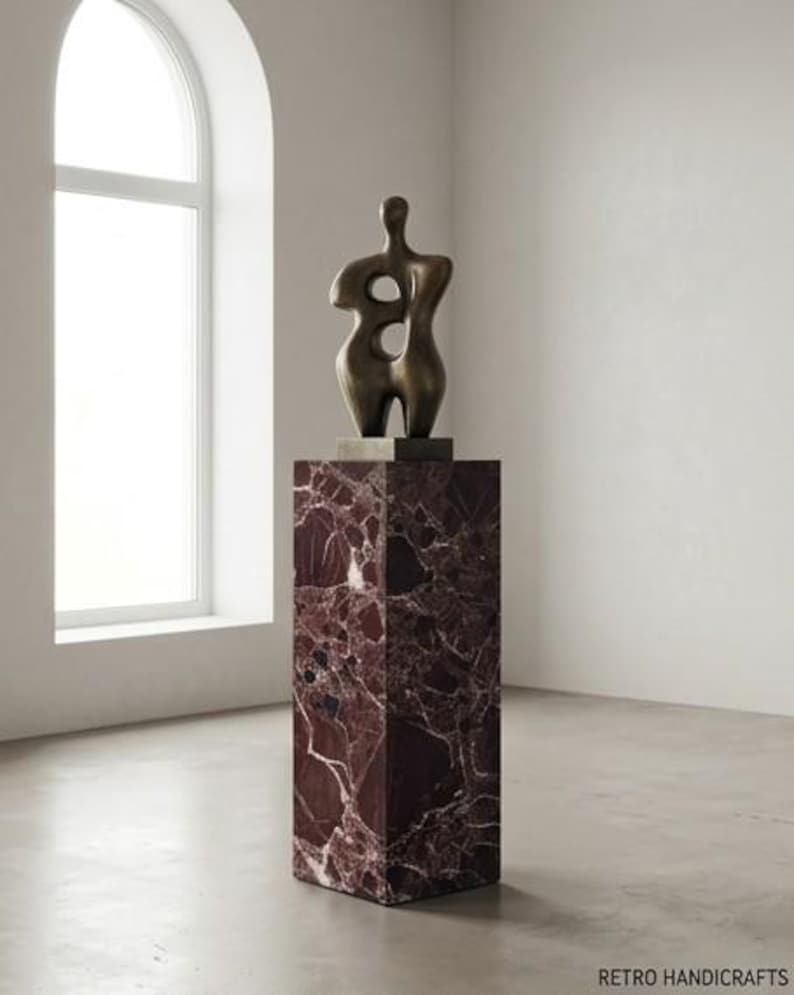May include: A bronze sculpture of a human form with two large openings, mounted on a rectangular, dark red marble pedestal. The sculpture is in a bright, minimalist room with a large arched window.