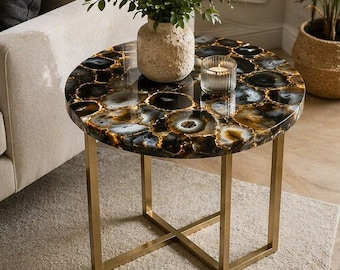 May include: Round side table with a black and gold agate stone top. The table has a gold metal base with an X-shaped support. A beige vase with greenery and a candle in a glass holder sit on the table.