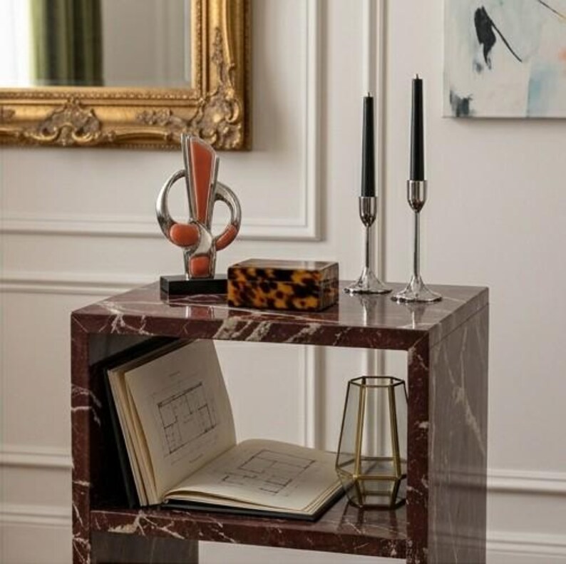 May include: A red marble side table with a decorative hourglass, a tortoiseshell box, two black candles in silver holders, and a gold geometric vase. An open book with architectural plans is on the lower shelf.