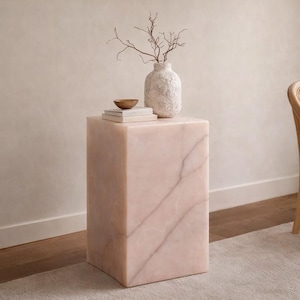 May include: A pink marble side table with a rectangular shape. A textured white vase with dried branches, a small gold bowl, and a stack of books are on top. A light-colored chair is to the right.