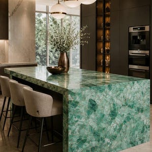 May include: A modern kitchen island with a vibrant green gemstone countertop. The island features a built-in seating area with three beige upholstered bar stools. Overhead, three pendant lights with gold accents illuminate the space. A vase with flowers and a bowl are on the countertop.