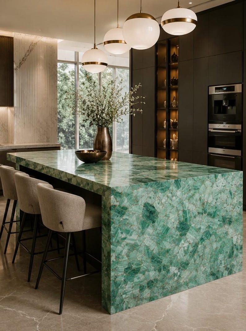 May include: A modern kitchen island with a vibrant green gemstone countertop. The island features a built-in seating area with three beige upholstered bar stools. Overhead, three pendant lights with gold accents illuminate the space. A vase with flowers and a bowl are on the countertop.