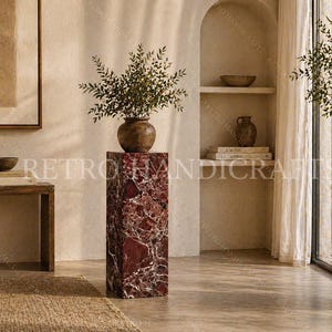 May include: A tall, rectangular pedestal made of red marble with white veining. A brown vase with green foliage sits atop the pedestal. The scene is set in a well-lit room with neutral tones and natural light.