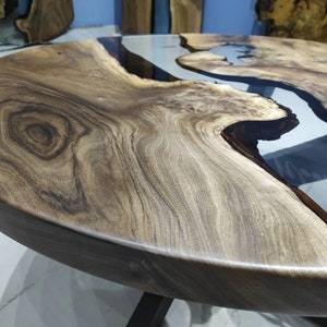 May include: A round wooden table with a natural wood grain finish. It has a clear epoxy resin river feature. The table is supported by black metal legs. The table's diameter is approximately 120 cm.