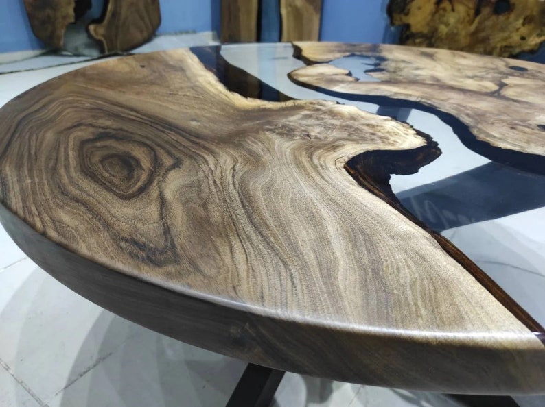 May include: A round wooden table with a natural wood grain finish. It has a clear epoxy resin river feature. The table is supported by black metal legs. The table's diameter is approximately 120 cm.
