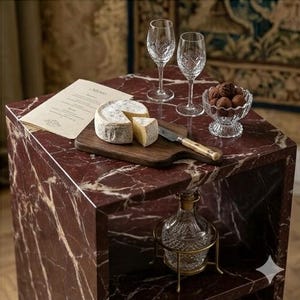 May include: A red marble side table with a cheese board, two wine glasses, and a small glass bowl of chocolate truffles. A decorative glass bottle with a gold metal stand sits below. A menu is on the table.